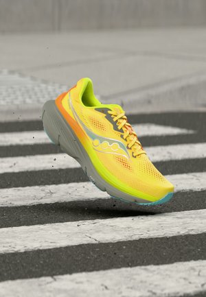 Világos sárga futócipő narancssárga és szürke részletekkel, levegőben egy fekete-fehér csíkozású gyalogátkelő felett, aszfalton.