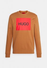 Brown sweatshirt with a crew neck, featuring a bold red rectangular patch with "HUGO" in black sans-serif font. Soft fabric texture.