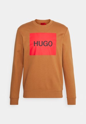 Bruine sweater met ronde hals, voorzien van een gedurfde rode rechthoekige patch met "HUGO" in zwarte sans-serif lettertype. Zachte stoftextuur.