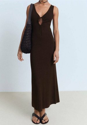 Femme portant une robe maxi marron foncé sans manches avec un détail noué à l'avant, un collier ras du cou noir, un sac à épaule noir et des sandales noires.