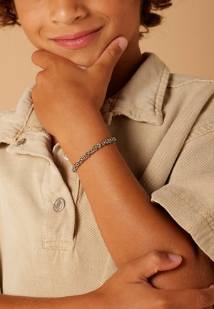 Enfant souriant aux cheveux bouclés portant un bracelet en chaîne argentée et une chemise beige à col, reposant le menton sur la main devant un fond neutre.