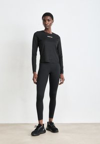 Een lange persoon staat met een zwart Calvin Klein lange mouw top, zwarte leggings en zwarte sneakers tegen een effen lichte achtergrond.