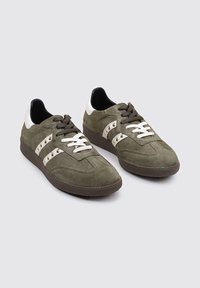 Zapatillas de ante verde con acentos blancos, superficie texturizada, puntera redonda, cordones emparejados y suela de goma marrón. Tres rayas a los lados.