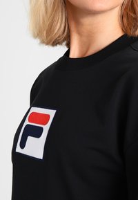 Mujer rubia vistiendo un sudadera negra con un gran parche del logo de FILA en rojo, blanco y azul marino en el pecho.