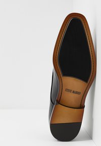 Semelle de chaussure habillée en cuir noir avec une finition lisse marron, comportant une empreinte subtile du logo. Forme arrondie avec une texture d'adhérence noire solide.