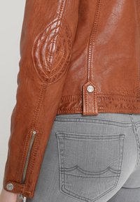 Mörkbrun läderjacka ärm med kviltat armbågsdetalj och dragkedja, i kombination med ljusgrå denimjeans där bakfickan syns.