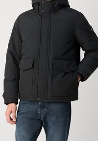 Musta toppatakki, jossa on huppu, kaksi etutaskua, napillinen suljenta ja teksturoitu kangas. Yksinkertainen muotoilu ja käytännölliset yksityiskohdat.