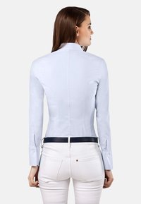 Chemise ajustée bleu clair avec un col haut et des manches longues, comportant des poignets boutonnés. Portée avec un pantalon blanc et une ceinture noire.