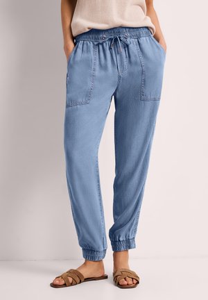 Person iført lyseblå elastiske taljedragter i denim med store forlommer og beige flade sandaler.