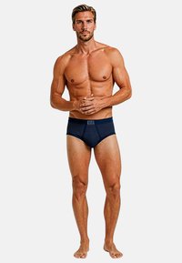 Slip a righe blu navy realizzati in tessuto morbido, con una vita standard e una vestibilità aderente, indossati da un modello maschile in piedi scalzo.