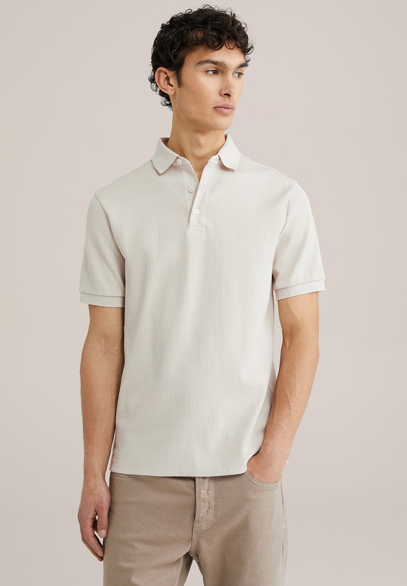 Gai pelēks ribots polo krekls ar plakanu apkakli, trīs pogu aizdari un īsām piedurknēm, kombinēts ar bēšiem biksēm. Minimālisma dizains.