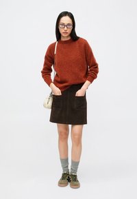 Pull en tricot couleur rouille, jupe en suède marron avec deux poches avant, chaussettes grises, baskets vertes et petit sac bandoulière blanc.