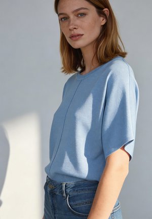 Frau mit schulterlangem hellbraunem Haar, die ein hellblaues Kurzarmtop und blaue Jeans trägt, vor einem schlichten hellen Hintergrund stehend.