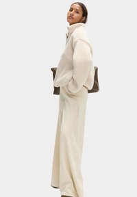 Kremowa polarowa kurtka z wysokim kołnierzem, luźny krój i długie rękawy; połączona z beżowymi spodniami o szerokich nogawkach; trzyma ciemno-brązową torbę.