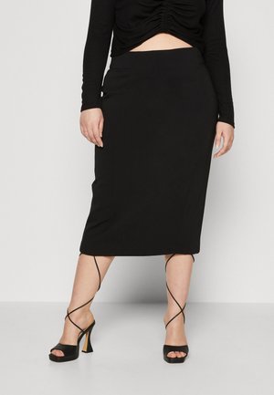 Femme portant une jupe midi noire, un haut à manches longues noir et des sandales à talons hauts noires avec de fines brides croisées à la cheville.