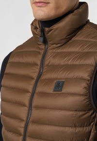 Braune Steppweste mit horizontalen Nähten, hohem Kragen und Reißverschluss vorne. Verfügt über ein kleines Logopatch auf der linken Brust. Glatte Textur.