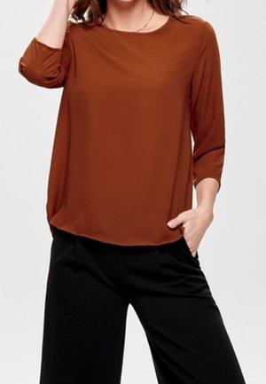 Vrouw draagt een roestkleurige blouse met lange mouwen en een zwarte broek, met één hand in haar zak en de andere hand raakt haar haar aan.