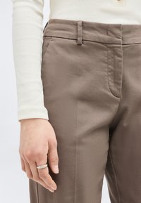 Κοντινό πλάνο χεριού ατόμου με ασημένιο στριφογυριστό δαχτυλίδι, που φορά καφέ παντελόνι και λευκό ριμπ πλεκτό μπλουζάκι με μακριά μανίκια.