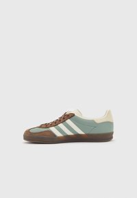 Zelená a hnědá sportovní obuv s bílými pruhy, z semišových a kožených materiálů, zaoblený špička, kontrastní gumová podrážka a hnědé tkaničky.