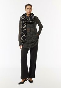 Black patterned long-sleeve top with a rounded hem, paired with sleek black trousers and a large patterned scarf in black and cream.
