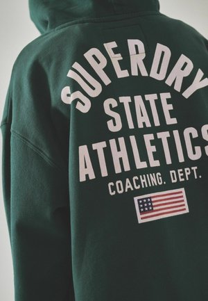 Zelená mikina s kapucňou s nápisom "SUPERDRY STATE ATHLETICS" vo veľkých bielych písmenách a grafikou americkej vlajky. Mäkká, štruktúrovaná tkanina.