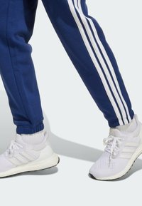 Pantalon de survêtement bleu avec trois rayures blanches sur le côté, associé à des chaussures de sport blanches avec une tige en maille et une semelle texturée.