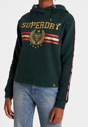 Person wearing a dark green Superdry hoodie with gold lettering and striped emblem, paired with light blue jeans.