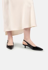 Picioare îmbrăcate în pantofi slingback cu toc mic și lucioși de culoare neagră, purtând o fustă midi din satin crem, pe un fundal alb simplu.