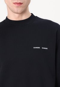 Sudadera de algodón negra con cuello acanalado, con el texto blanco "SAM SøE" bordado en el pecho en dos líneas.