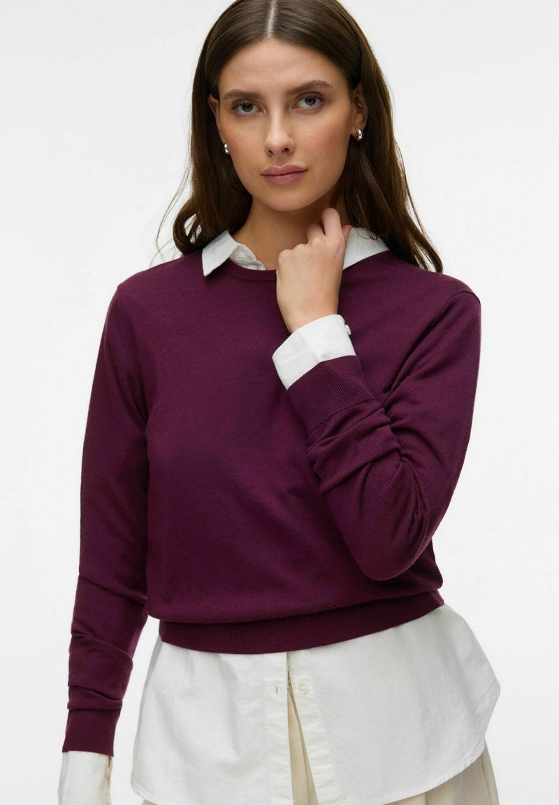 Femme portant un pull violet par-dessus une chemise blanche à col, touchant son cou, sur un fond blanc uni.