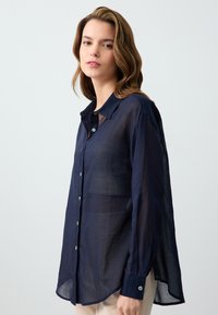 Navy blue, sheer button-up shirt with a collar, long sleeves, and a relaxed fit. Features shiny buttons and a side slit design.
