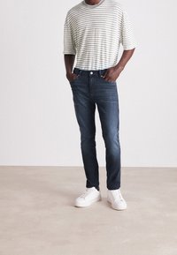 Hombre vestido con una camiseta de rayas blancas y grises, jeans azul oscuro y zapatillas blancas, de pie con las manos en los bolsillos sobre un suelo beige.