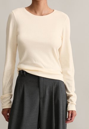 Femme portant un haut moulant à manches longues crème rentré dans un pantalon large gris foncé taille haute sur un fond uni.