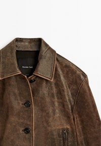 Giacca in pelle marrone con chiusura frontale a bottoni, grande colletto e tasca laterale con zip. Presenta una superficie texturizzata e dettagli di cucitura in contrasto.