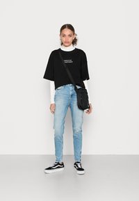 T-shirt oversized preta com texto branco, sobreposta a uma camisa de manga longa branca; jeans azul claro; mochila preta; ténis preto e branco.