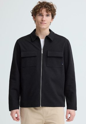 Ung mand med krøllet hår iført en sort lynlåstrøje med to forlommer med klap og beige bukser, stående foran en ensfarvet baggrund.