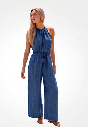 Frau trägt ärmellosen blauen Halter-Jumpsuit mit Kordelzug in der Taille und weiten Beinen, steht mit dem Kopf zur Seite gewandt und trägt Sandalen.