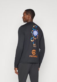 Μαύρο πουκάμισο με μακριά μανίκια που διαθέτει πολύχρωμο γραφιστικό σχέδιο στο πίσω μέρος, το οποίο περιλαμβάνει κυκλικά λογότυπα και ένα σχέδιο που μοιάζει με φίδι.