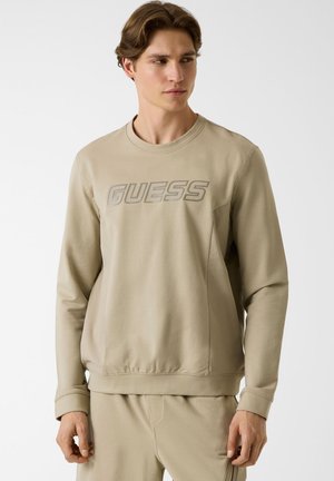 Jeune homme portant un sweat-shirt beige et un pantalon assorti, le sweat-shirt porte le logo "GUESS" sur la poitrine, debout devant un fond uni.