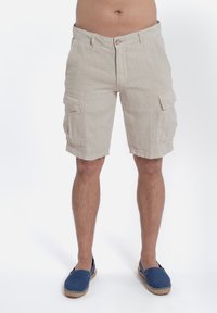 Shorts cargo in lino beige con due tasche laterali, chiusura con bottone e zip, e orlo non rifinito. Indossati con espadrillas blu.