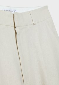 Beige-linjakankaiset housut ovat litteällä etuosalla, hienovaraisilla laskoksilla ja sileällä vyötärönauhalla. Saumoissa oleva ommelkoristelu lisää tekstuuria.