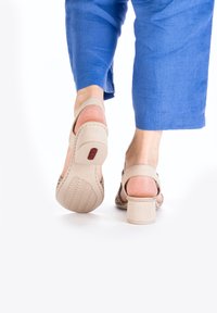 Sandales à talons beiges avec une semelle texturée et un dos ouvert, dotées d'une large lanière et d'une bande de cheville ajustable, portées avec un pantalon cropped bleu.