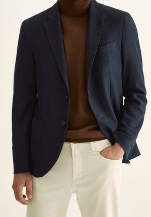 Mężczyzna w granatowej marynarce nałożonej na brązowy sweter z golfem i białe spodnie, jedna ręka w kieszeni, druga swobodnie opadnięta wzdłuż ciała.