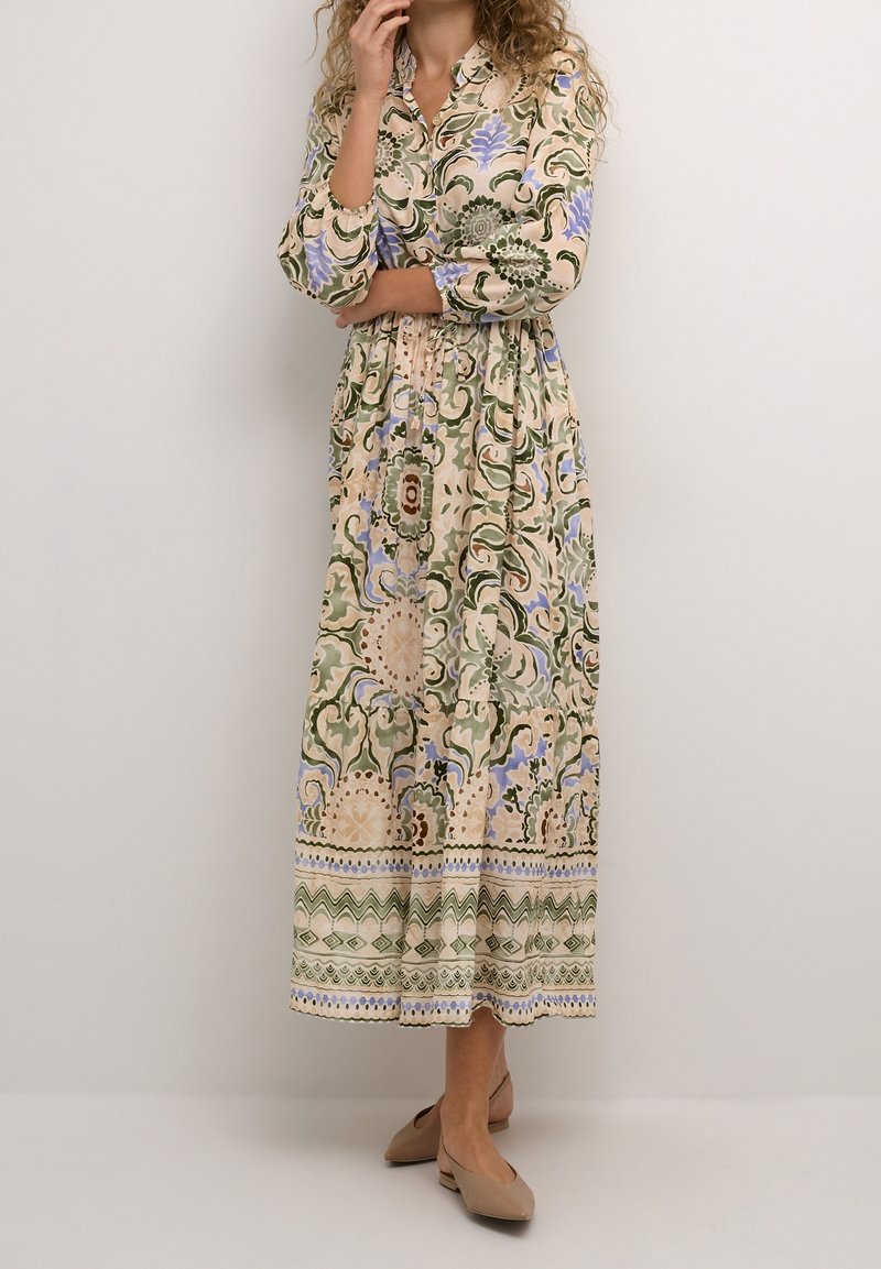 Femme portant une longue robe à motifs beige, verte et bleu clair à manches longues, debout, une main touchant son visage et l'autre croisée.