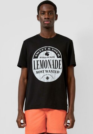 Jonge man met een zwart T-shirt met witte tekst "Sweet & Sour Hand Picked Lemonade Most Wanted" en oranje korte broek, staand tegen een grijze achtergrond.