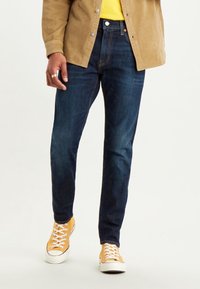 Jeans en denim bleu foncé coupe slim, associés à des baskets jaunes et une veste en velours côtelé beige portée sur une chemise jaune.