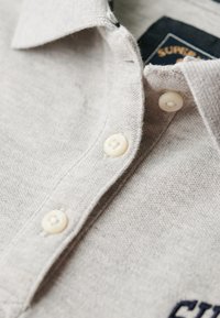 Polo de color gris claro con una tela texturizada, que presenta una tapeta con botones, cuatro botones, y un detalle bordado en color marino en la parte delantera.