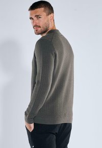 Pullover in Olivgrün, strukturiertes Strickmuster, lange Ärmel, gerippte Bündchen und Saum. Modell von hinten auf neutralem Hintergrund gezeigt.