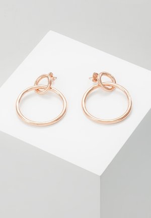 Boucles d'oreilles créoles en or rose avec une petite boucle attachée en haut, présentées en biais sur un socle blanc contre un fond gris clair.