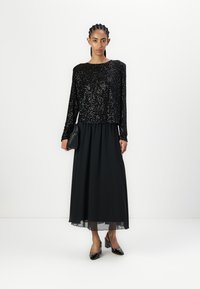 Čierny top s flitrami a dlhým rukávom v kombinácii s prúdiacou čiernou maxi sukňou. Model nosí čierne lakované lodičky a drží malú čiernu kabelku.